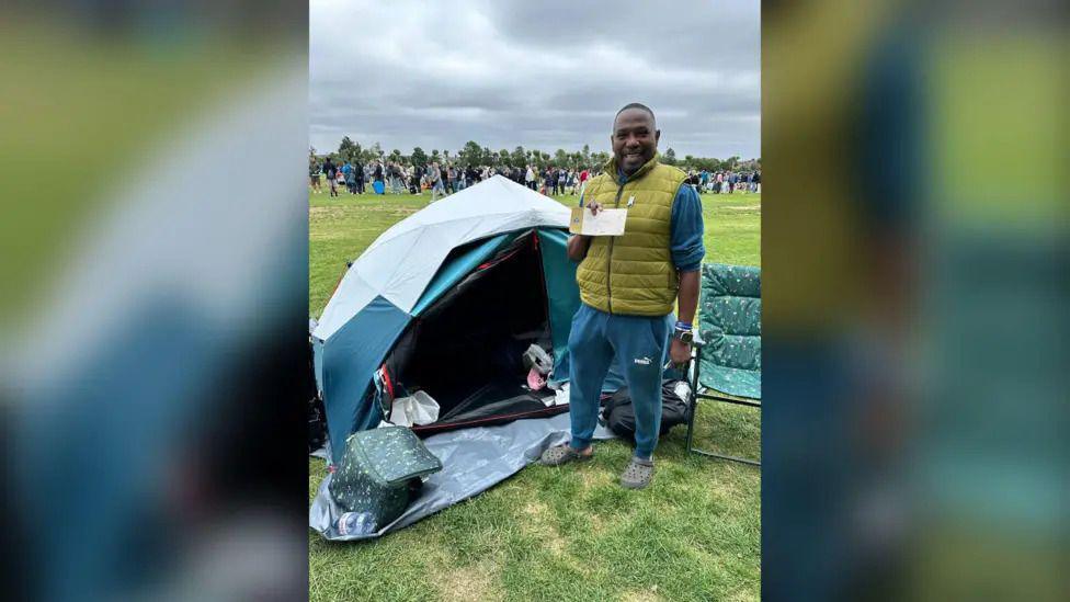 A man standing in a field next to a tent. He is smiling an holding a ticket.