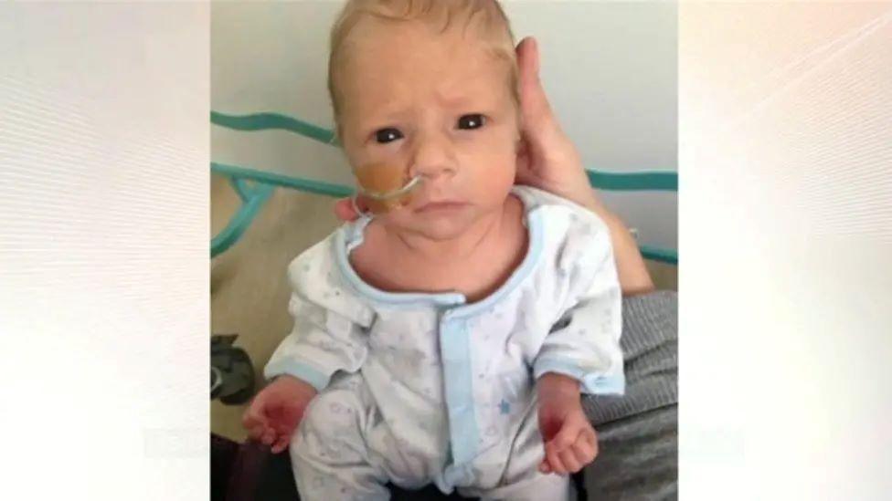 A baby in a white and blue baby grow is held up to the camera. He has a nasal tube. 