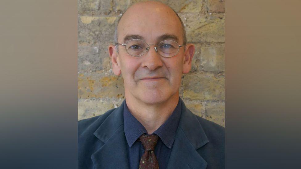 Head and shoulders image of Ben Crystall in front of a brick wall. He is wearing glasses, a dark blue jacket and shirt and a brown tie.