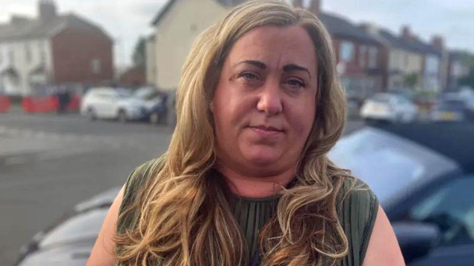 Leanne Hassan, with long blonde hair and wearing a green vest top, stands in front of a parked car and a row of terraced houses