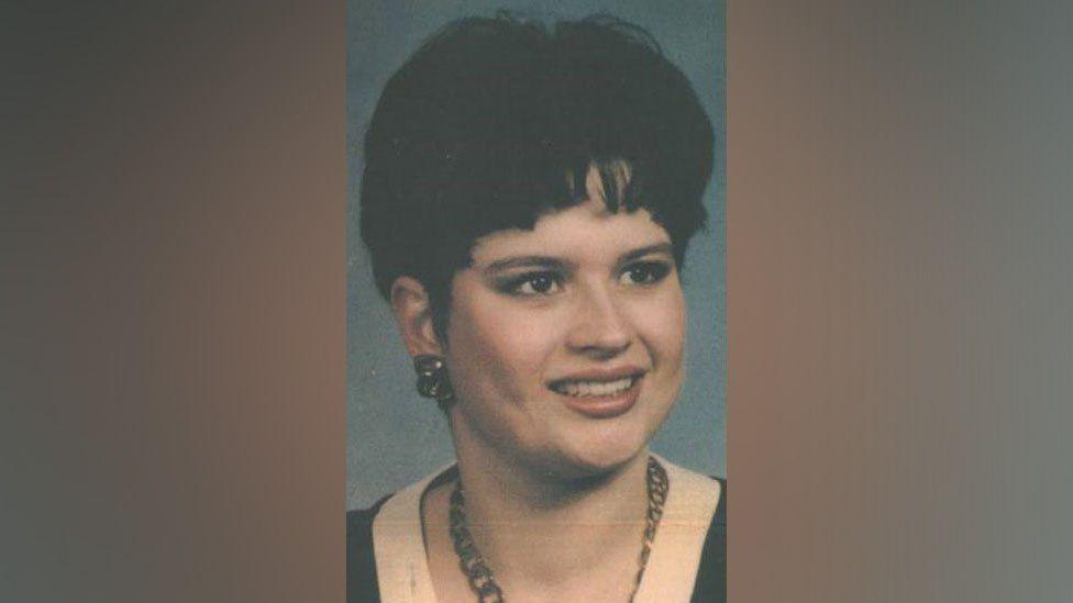 A portrait shot of Karen Hales, who has dark hair and earrings and is wearing a necklace and a black top with a white top underneath. 