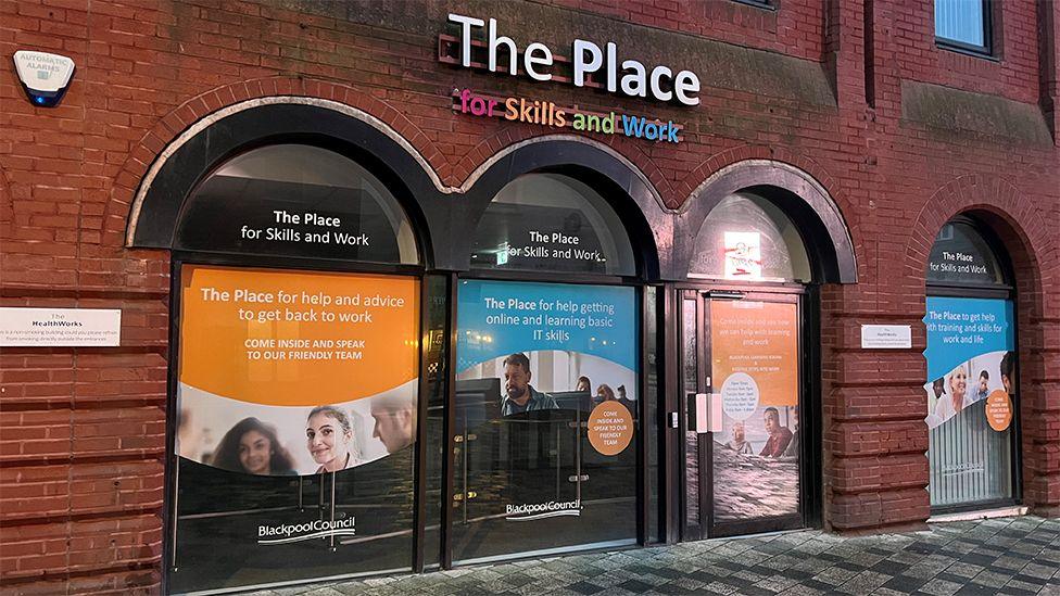 The safe space project building on Clifton Street. It is a red brick building with four big arched windows with posters offering help and advice and a sign above them saying The Place for skills and work