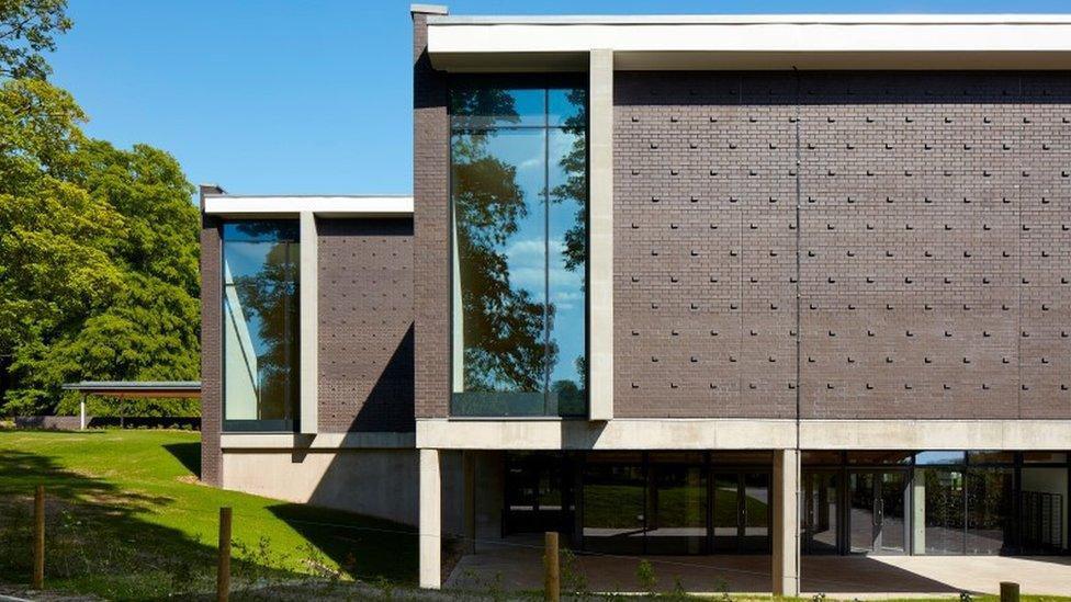 An exterior shot of the St Fagans visitor centre entrance. The building sits on the side of small slope of grass. It is brown and beige with large glass windows. 