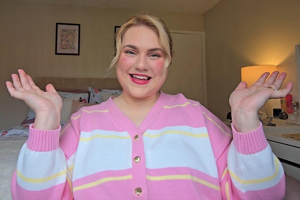 Isobel Guffick, a blonde woman in a pink, white and yellow cardigan, sits in a bedroom and smiles at the camera with her hands in the air