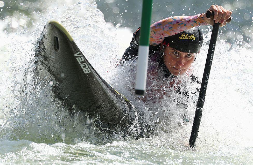Elena Lilik competes in the Women’s Canoe Slalom heats at the Slalom World Championships in Penrith, Australia. She maneuvers a black Vajda canoe through churning whitewater, gripping a paddle while passing a green slalom pole as water splashes forcefully around her.