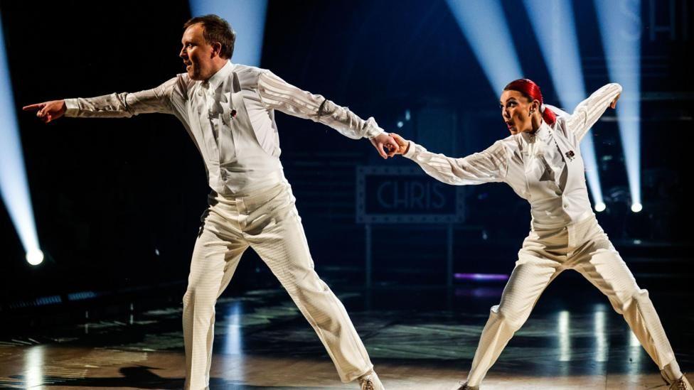 Chris McCausland and Dianne Buswell performing on Strictly in white suits
