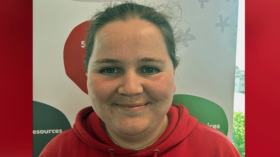 Joanne wears a red hooded top and smiles, looking at the camera. Her dark brown hair is tied back.