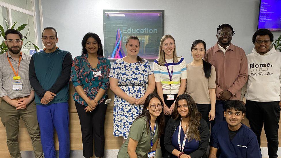 Eleven of the trainee doctors, dressed in casual clothing and smiling in front of a reception desk at the hospital