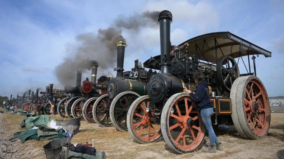 Long line of steam engines with black smoke billowing into the sky they are black , some with red painted spokes on their wheels a person leans against the wheel of the first engine