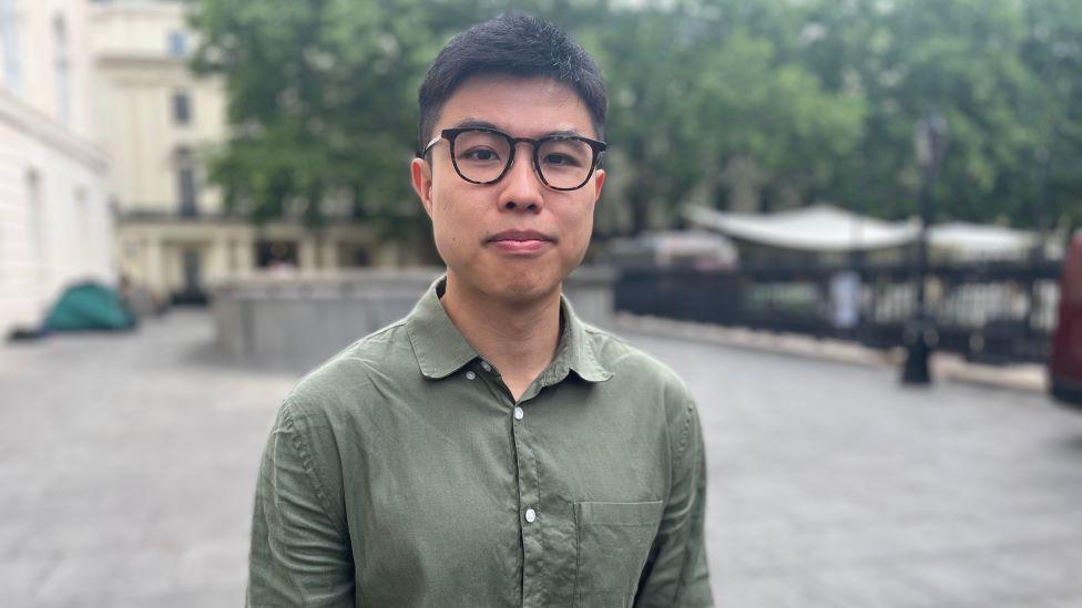 A man, stood outside by paved floor, is wearing a khaki shirt and glasses. He has short, dark hair and is looking at the camera