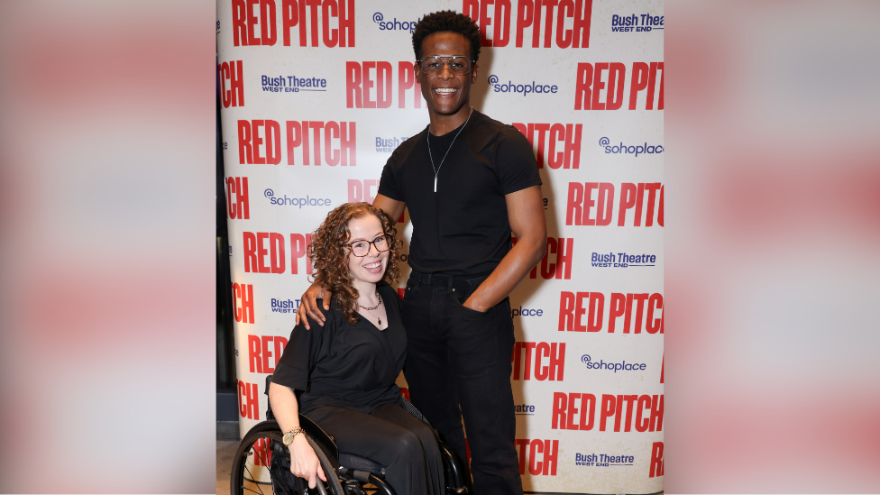 Amy Trigg and Jordan Benjamin. Both are wearing black and smiling at the camera. They are standing in front of a promotional board at the Soho Place theatre in London.