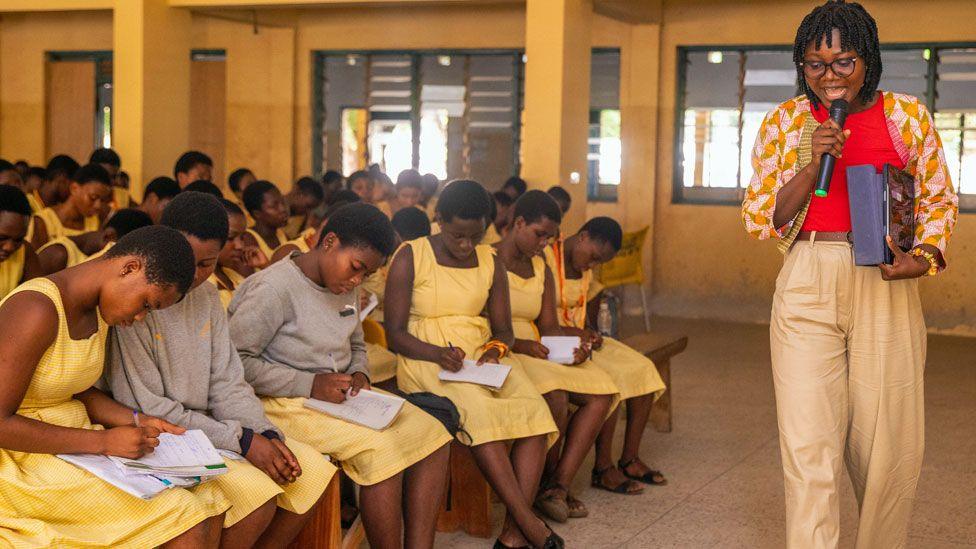 Dr Angela Tabiri walks and talks holding a microphone during a class and Innovate Her outreach event at Accra Girls Senior High School. The students, all in yellow dress uniforms, take notes as they sit on benches in a classroom.