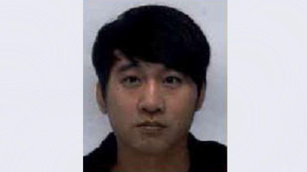 A grainy police mugshot of Zhaoqian Yu wearing a black hoodie and standing against a grey background. He has brown eyes and black hair styled in a thick sweeping fringe.