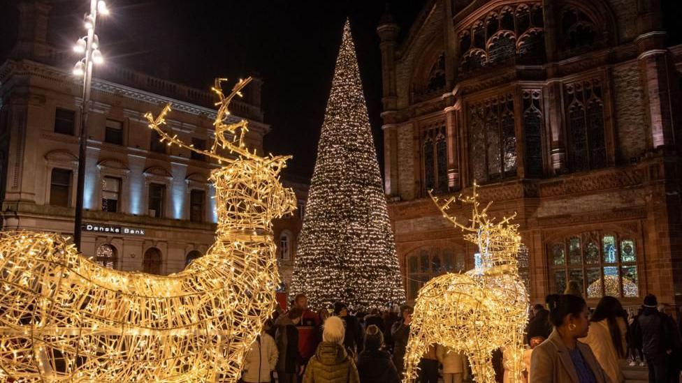 Two illuminated reindeer decorations are positioned behind a civic Christmas tree in Derry which is also lit up. The Guildhall is in the background. A number of people are gathered around the decorations.