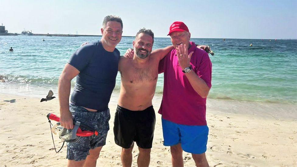 Christopher Brightmore is wearing a purple shirt, blue shorts and red cap. He is standing on a sandy beach with two men and he is on the right with his left-hand raised up towards his face. There is a ring on his middle finger and a black-strapped watch on his wrist. Behind him is turquoise-coloured water lapping against the shore. Christopher's right arm is across the shoulder of the man standing in the middle, who is wearing a pair of black shorts. The man in the middle is standing with both of his arms stretched across the shoulders of the two men either side of him. Diver Adam is standing on the right and he is wearing a navy t-shirt and navy patterned thigh-length shorts. He is holding a red and black snorkel in his right hand.