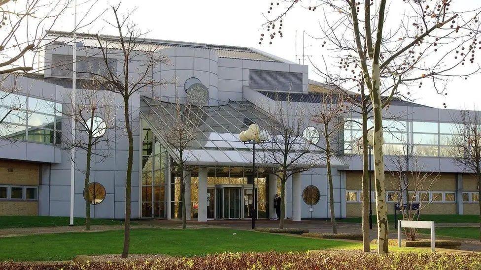The outside of Woolwich Crown Court, a modern building with a glass sloping entrance roof and a lawn with trees in front of it.