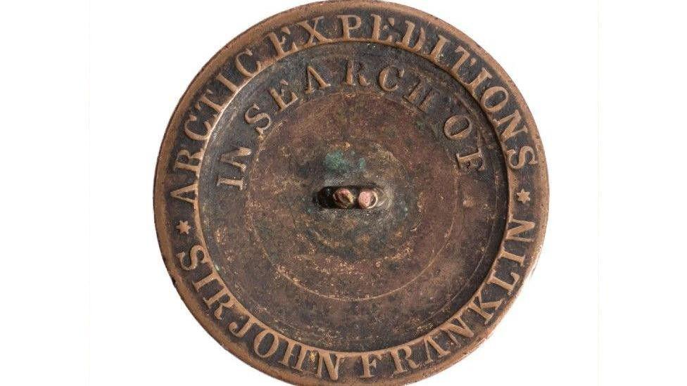 The button. It is brown. It says 1852 on it and has writing going along various bits of it. It is on a white background.