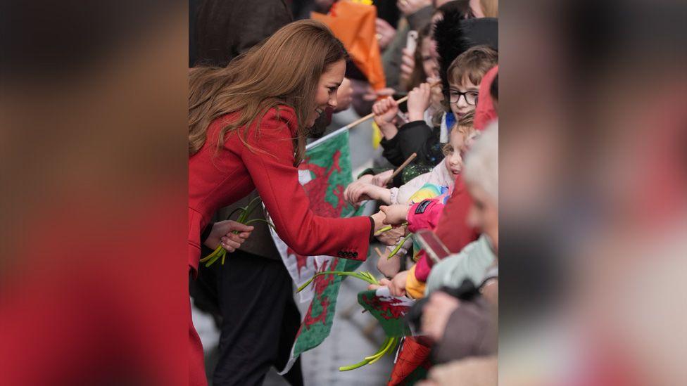 Catherine meets children. She bends down talking to them, many holding bunches of daffodils and Welsh flags.