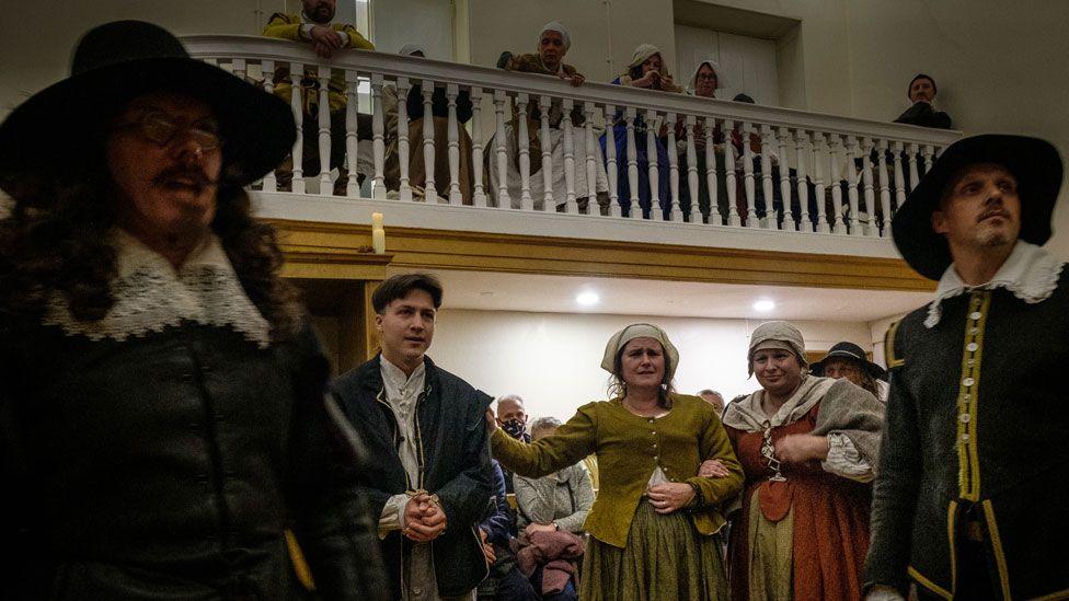 A  re-enactment of the Huntingdon witch trial in an 18th Century courthouse. Three men and two women in mid 17th Century dress and two women are standing and above them is a balcony where other people in mid-17th Century dress can be seen. Two of the men are wearing broad brim black hats and dark clothing with wide lace-collars above their jackets. The women are in russet-toned dressed with white caps on their heads. 