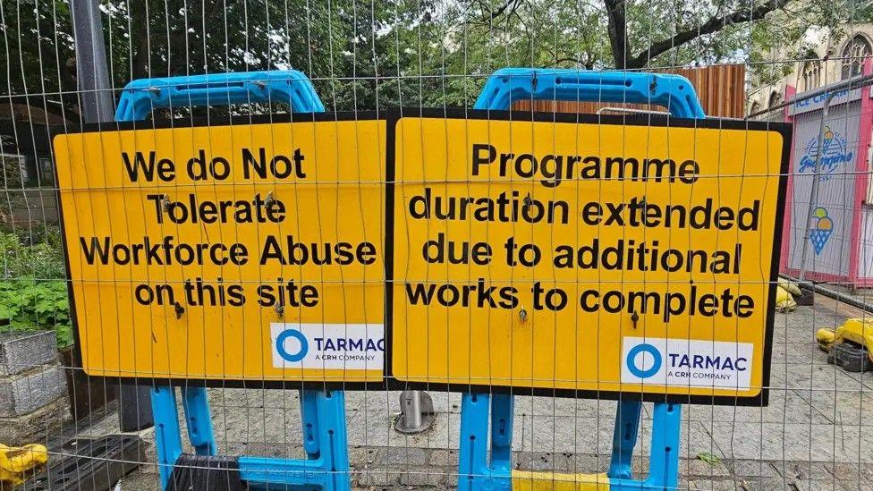 Two yellow work signs. The sign on the left says, "we do not tolerate workforce abuse on this site". The sign on the right says, "programme duration extended due to additional works to complete"