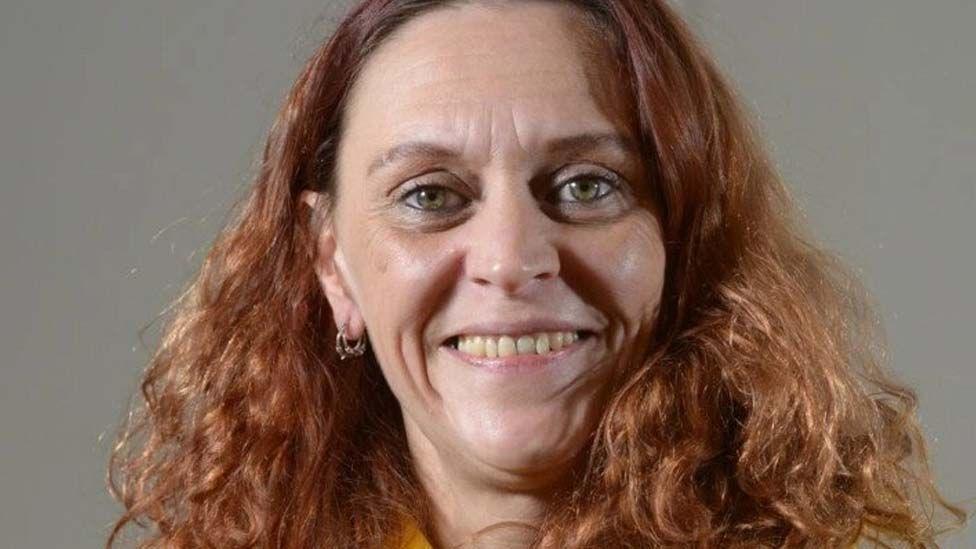 Head shot of Jenny Hurley, she is smiling in front of a grey background and has long curly red hair