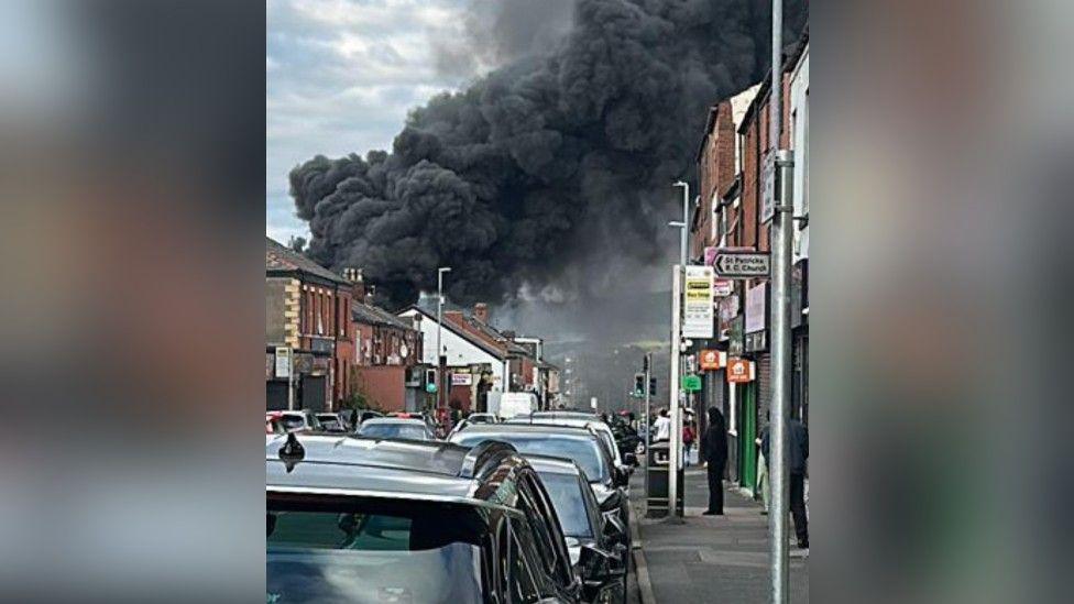 Black smoke pours into the sky from above a row of homes, with onlookers stood watching on from the pavement on the roadside. 