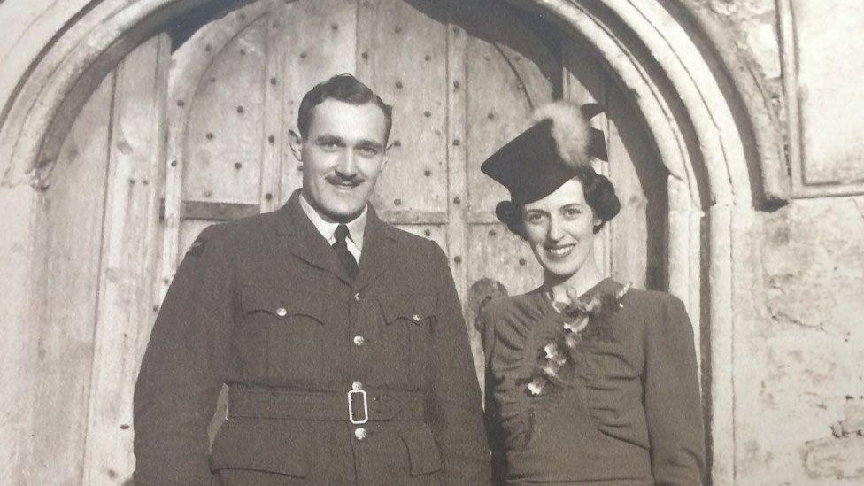 A sepia-toned black and white image showing Cuthbert "Cubby" Lander and his wife Eileen. They are standing outside a stone-built church's wooden door, having just got married. It is World War Two and Cubby is shown wearing an RAF uniform. He has short hair, a short moustache and is smiling broadly. On the left is Eileen, wearing a dark hat, with dark hair pulled back and a short dress. She is also smiling broadly. 