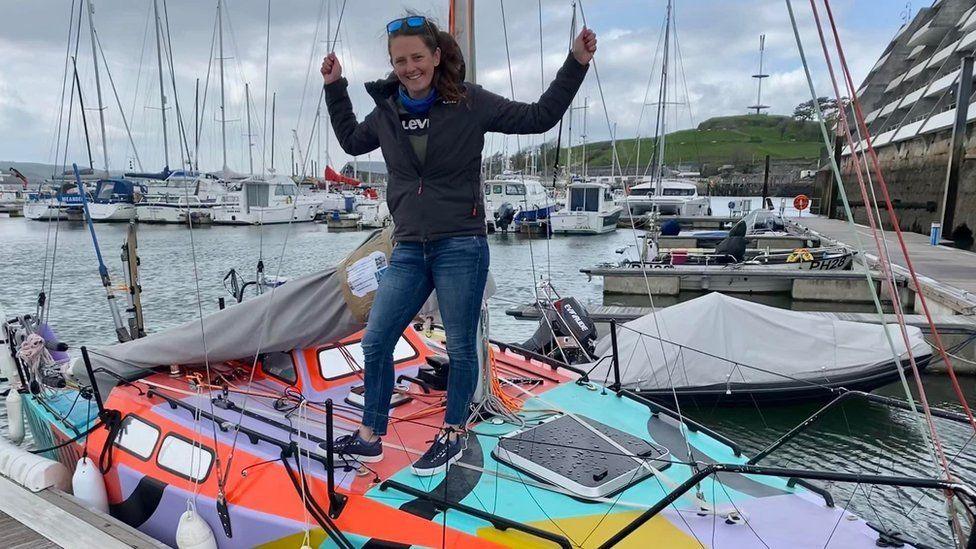 A picture showing Jasmine Harrison who is smiling and wearing jeans, trainers and a black jacket with sunglasses on the top of her head. She is stood on a sailing yacht which is on the water. In the background are several other boats. 