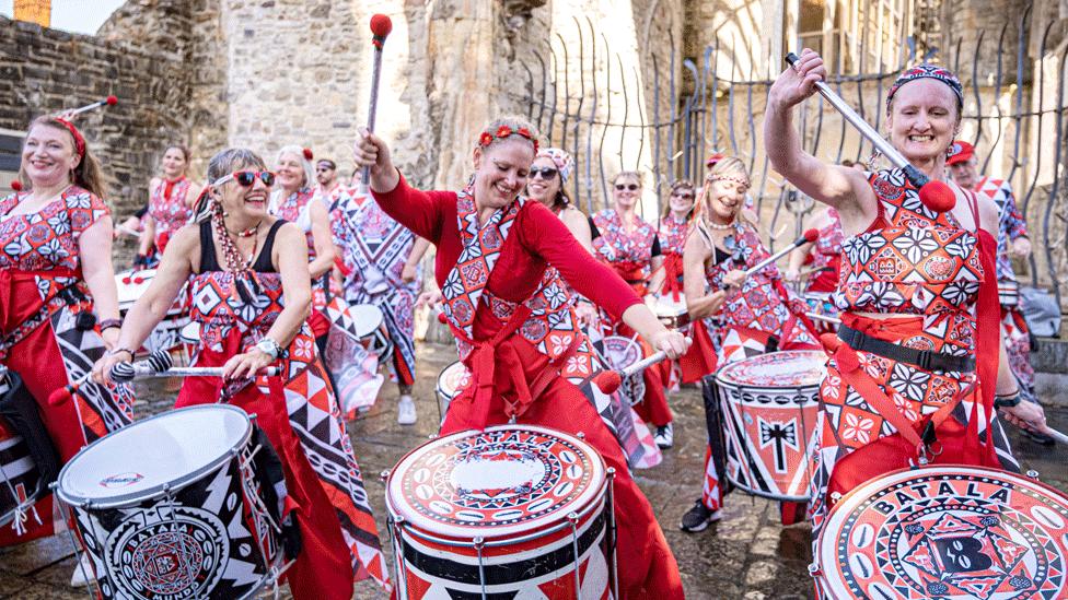 Women in traditional dresses drum a samba outside a church.