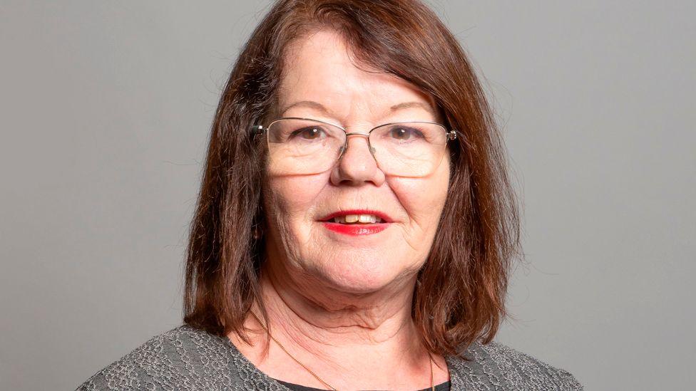 Head and shoulders image of Kate Hollern against a grey background. She has shoulder length brown hair and is wearing glasses and smiling