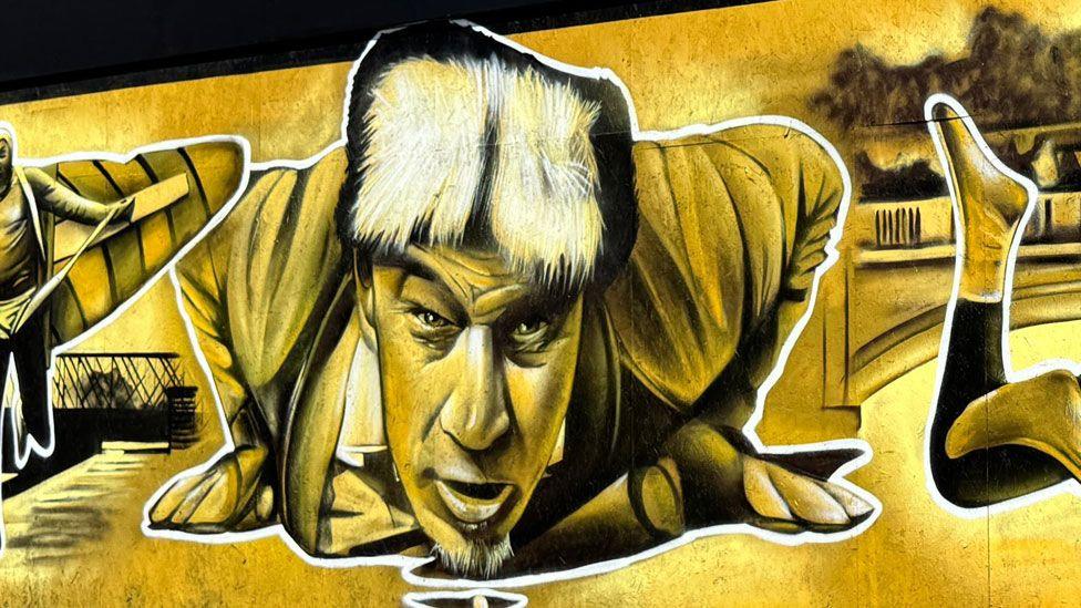 A close up of part of a mural, painted in yellow, black and white, showing Walter Cornelius, face on and flat on his stomach with his hands beneath his shoulders. His head is raised and he is looking forward and in front of him on the ground is a pea.