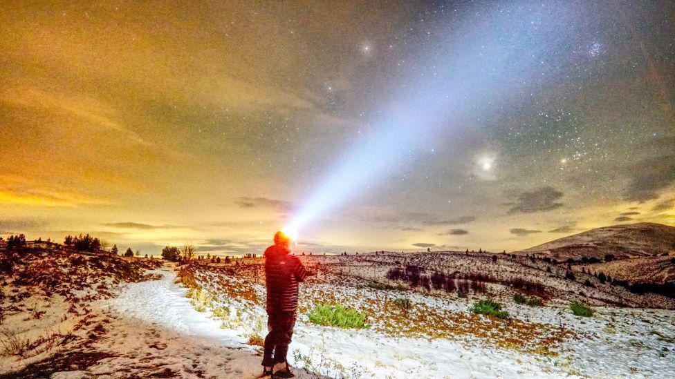 Man in dark clothes on snowy hillside wearing head torch shining into a starry night sky.