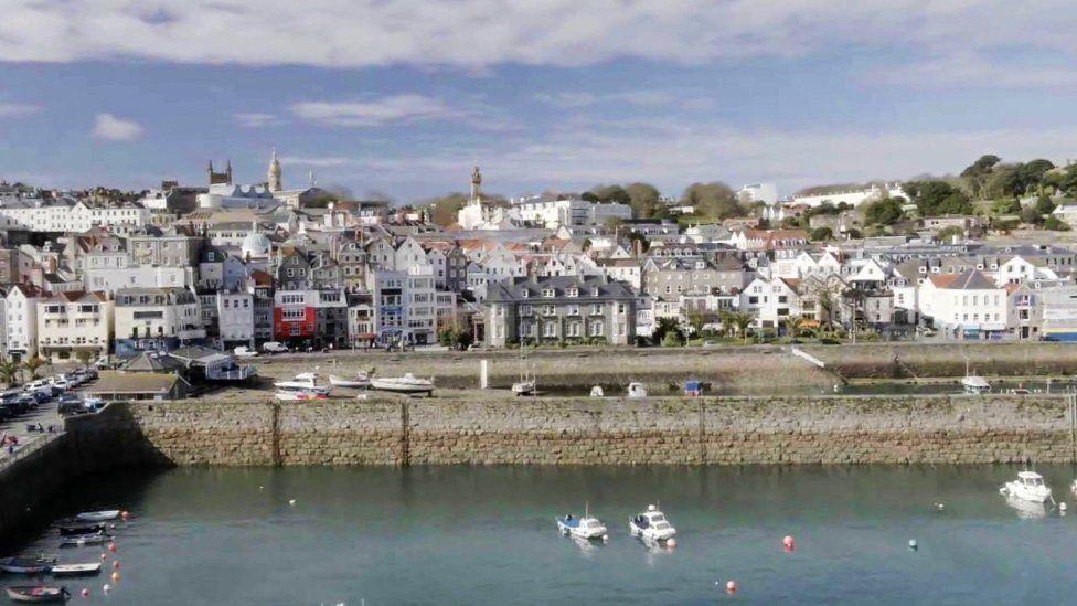 Guernsey harbour with water in the forefront and a number of houses in the back, with blue sky at the top 