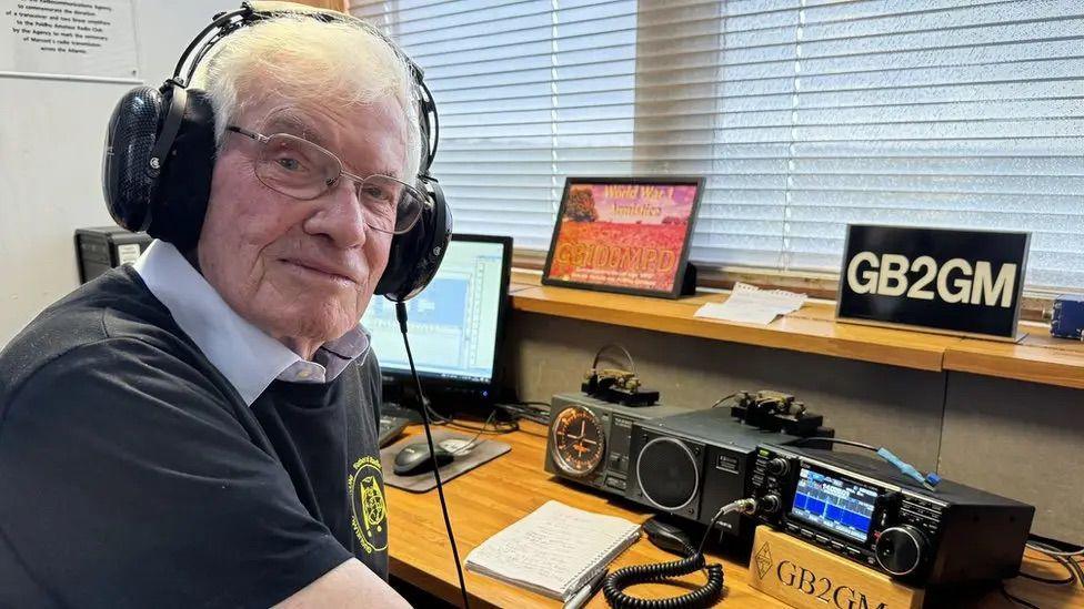 David Barlow, a radio enthusiast, wearing headphones and a navy blue shirt and glasses. There is radio equipment on the table in front of him.