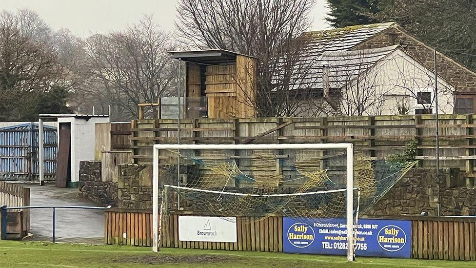 The goalpost at Barnoldswick Town FC. In the background is a wooden fence with the wooden clad football stand peering over the top