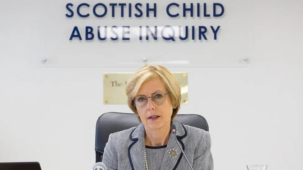 Lady Smith is sitting under a blue-lettered sign on a grey wall saying 'Scottish Child Abuse Inquiry'. She is wearing a grey suit with blue piping on the collar. She has a pearl necklace.