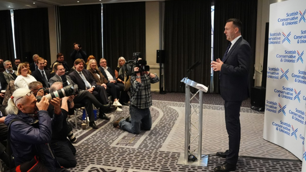 Russell Findlay stands at a podium wearing a black suit as party officials and media camera crews look on.