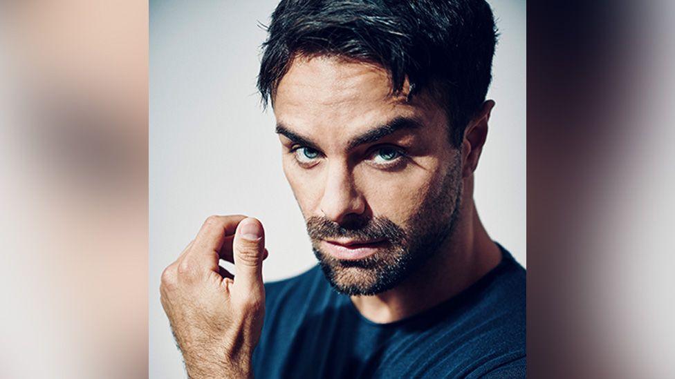 A headshot of a man taken against a white background. He's got jet black hair, cut short and neat, and a well-manicured beard. He's looking straight down the camera lens, highlighting his piercing blue eyes. The lighting creates shadow on the left side of his face, creating a sense of mystery. 