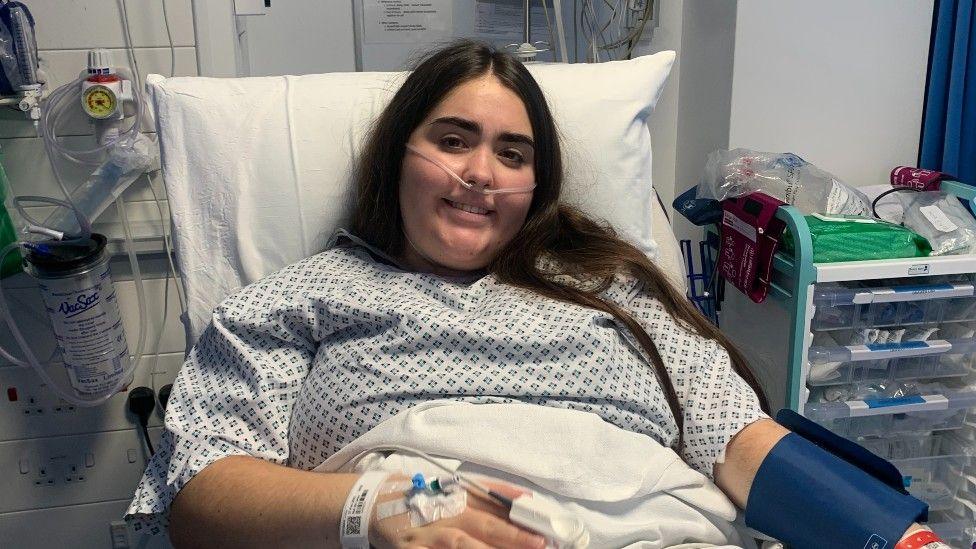 A young woman with long dark hair wearing a hospital gown lying in a hospital bed, with an intravenous canula in her hand and a nasal canula in her nose