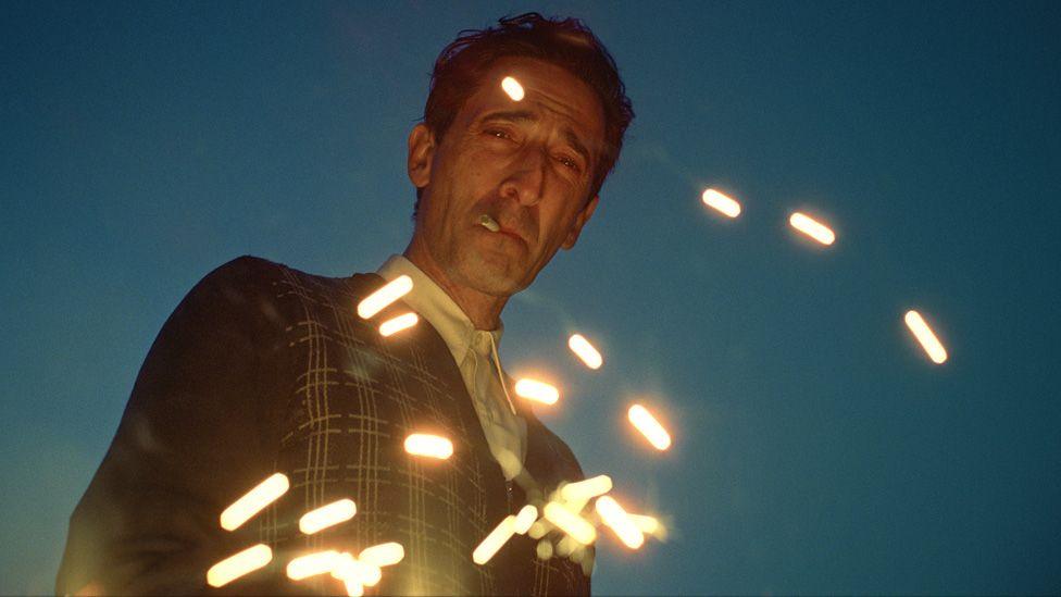 Adrien Brody smoking in the twilight apparently looking into a fire as sparks fly up in The Brutalist