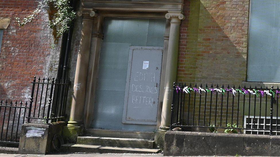 Close up of the doorway of the red brick house, which is boarded up with metal. There is graffiti on the door which says "Edith deserves better"