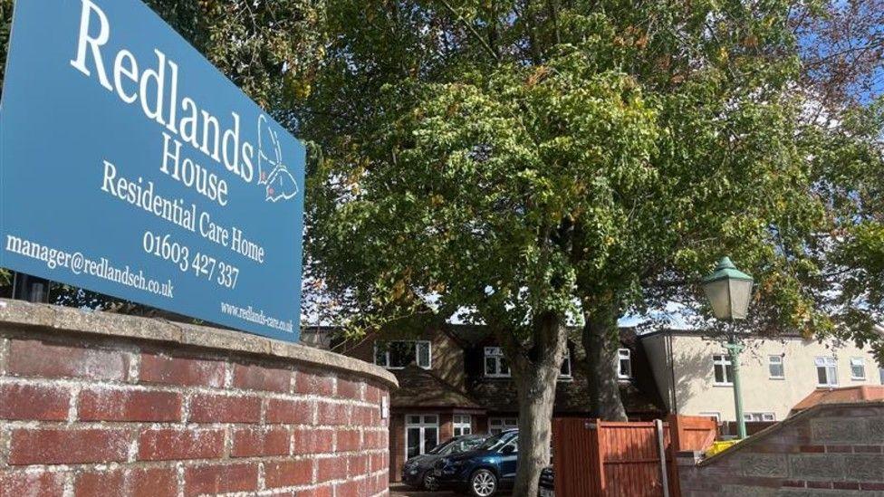 View from the street of the front outside of Redlands House care home. There is a red brick wall with a blue sign attached with the name on. A large tree is in front of the red brick building and two cars are parked outside. 