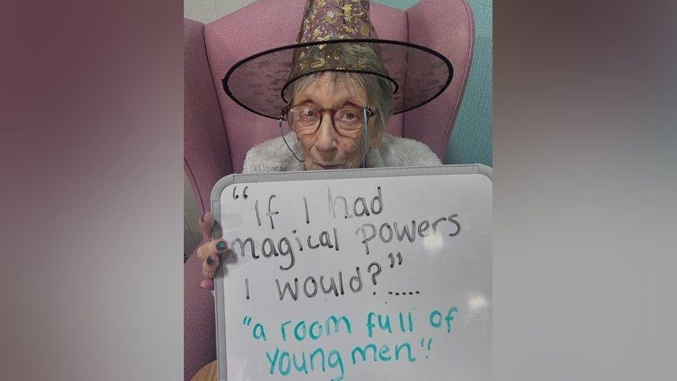 An elderly woman sitting in a pink chair wearing glasses and a witches hat holding up a sign which reads "if I had magical powers I would?" with the answer "a room full of young men".