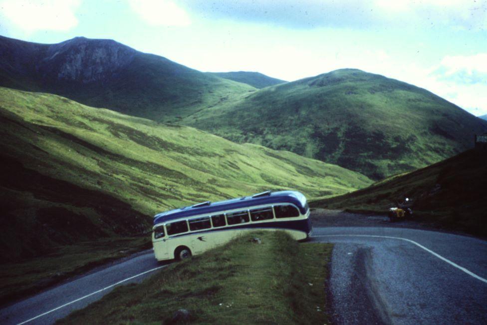 Old cream and blue-coloured single-decker bus negotiating a winding hillside road with green slopes in the background.