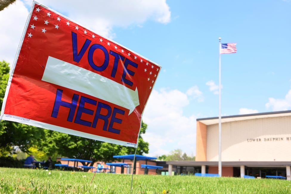 Sign saying 'Vote here' on a lawn in front of a building