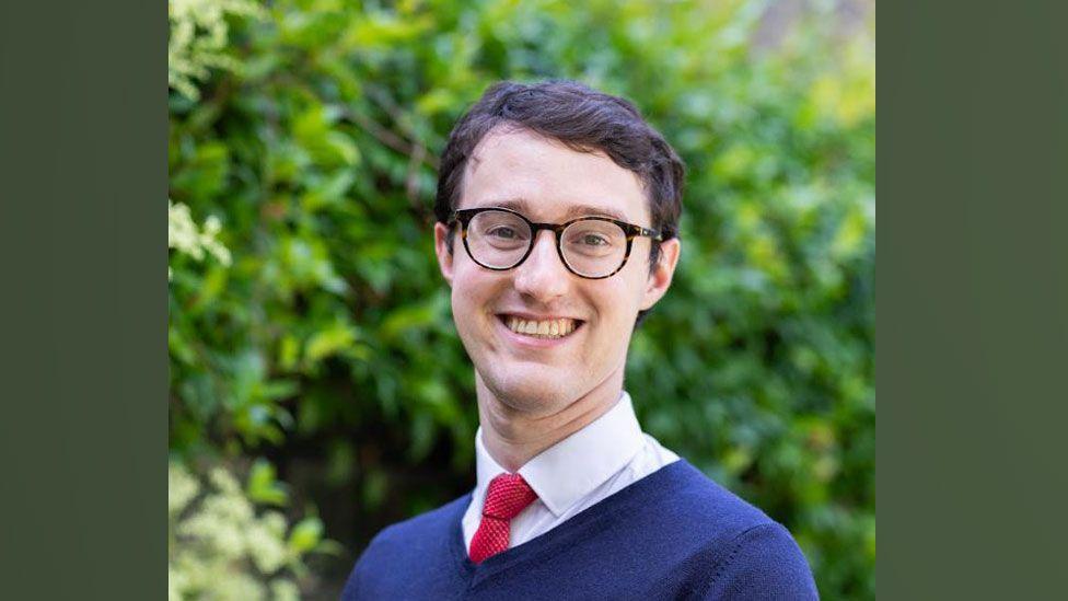 Dr Eamonn O'Keeffe, who has short dark hair and dark framed glasses. He is wearing a blue v-necked jumper over a white shirt and red tie. He is smiling towards the camera and behind him are the blurred-out green leaves of a tree or shrub.