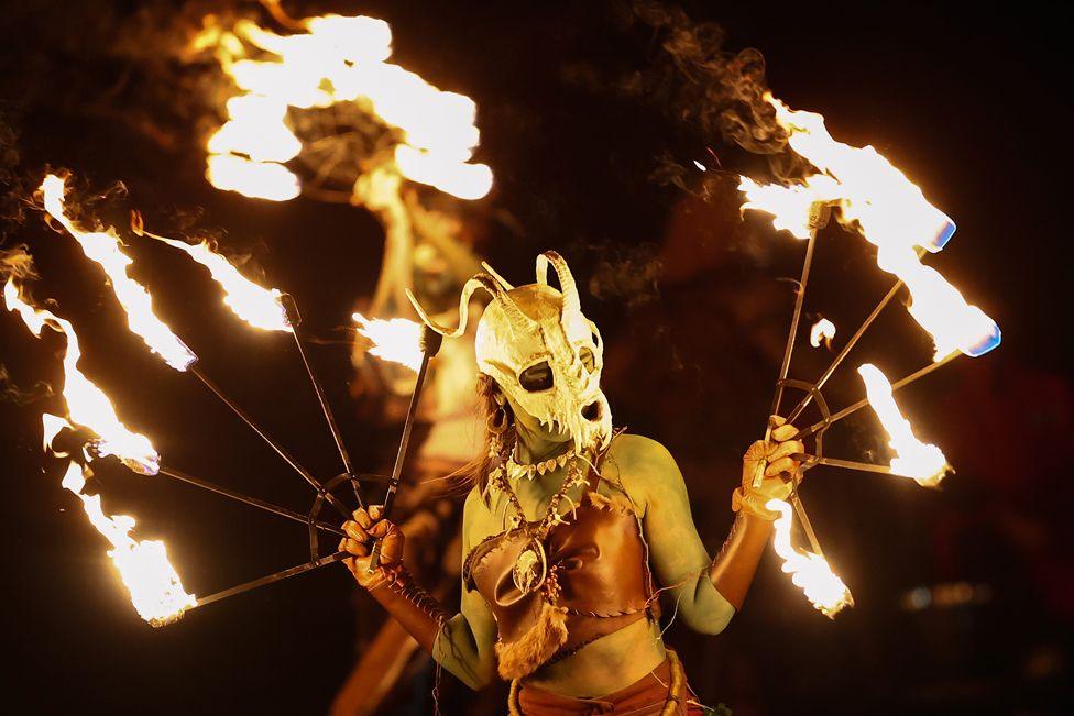 A costumed woman wearing a mask performs a fire-dance. She is painted green and holds a metal metal frame of five flaming torches in each hand