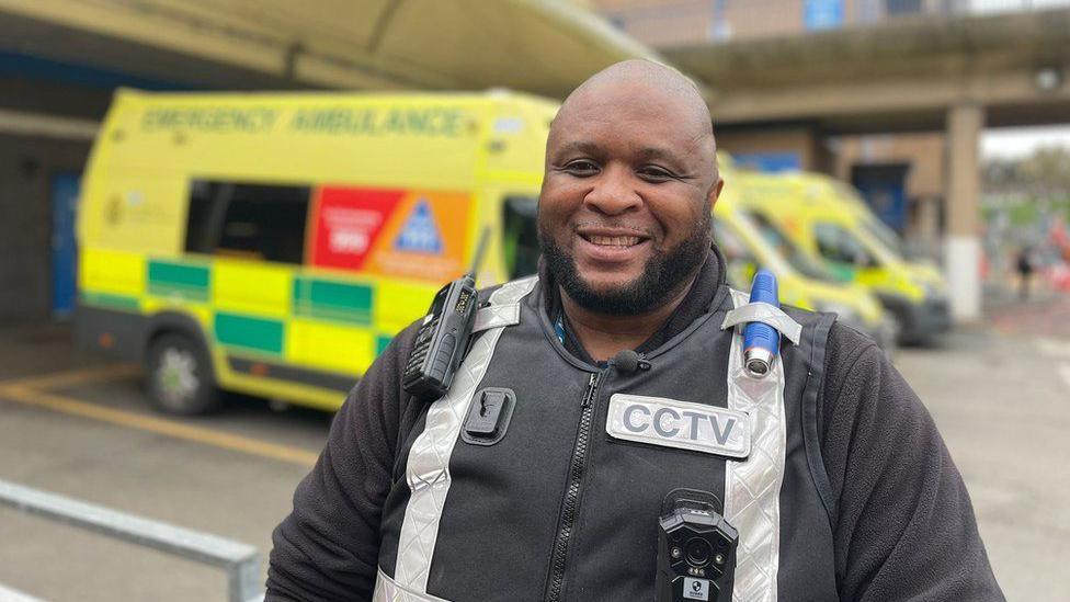 Leston Scafe, smiling at the camera, stood outside the hospital, wearing his 
 uniform, with ambulances in the background.