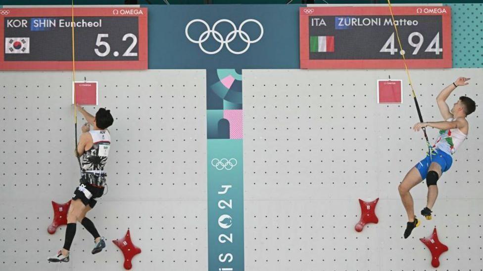 Two climbers with ropes attached to their waists, hang at the top of a white speed climbing wall with red holds.