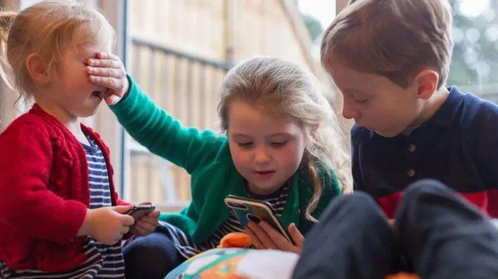 Three young children sit in a classroom, there are two little girls both with blonde hair sat on the left who is holding a phone in her hands and in the middle who is also holding a phone but she has one hand over the other little girl's eyes, and a young boy with light brown hair on the right, who is looking at the girl in the middle's phone